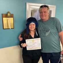 Susan holding certificate beside husband after completing chemotherapy treatment, smiling and wearing cold cap head covering