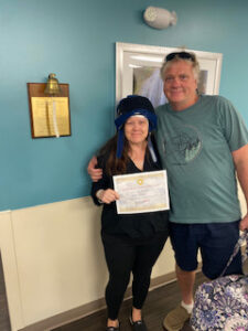 Susan holding certificate beside her husband after completing chemotherapy treatment, smiling and wearing cold cap head covering
